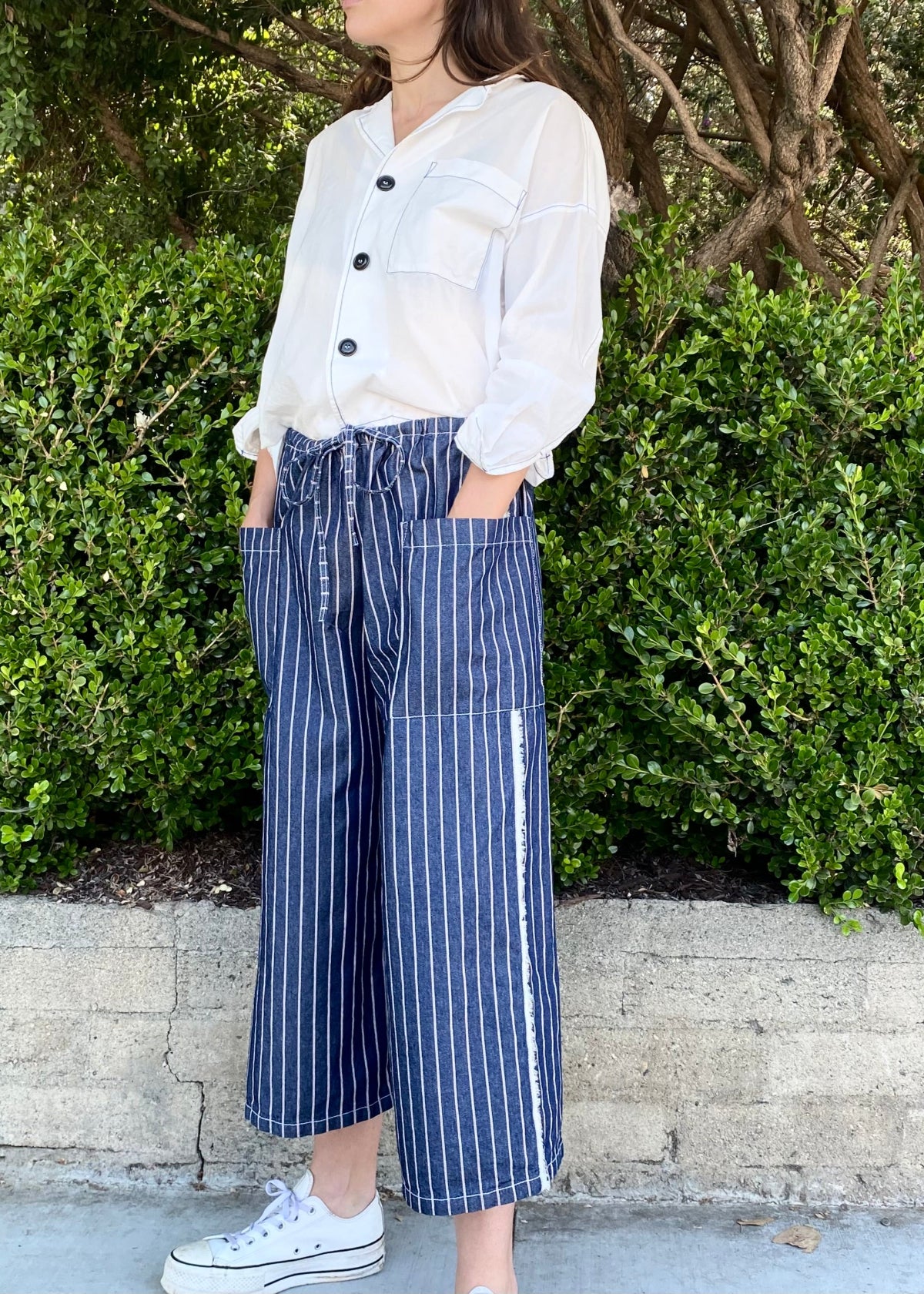 blue and white striped denim cropped drawstring pants with white button down top. Hands in pockets