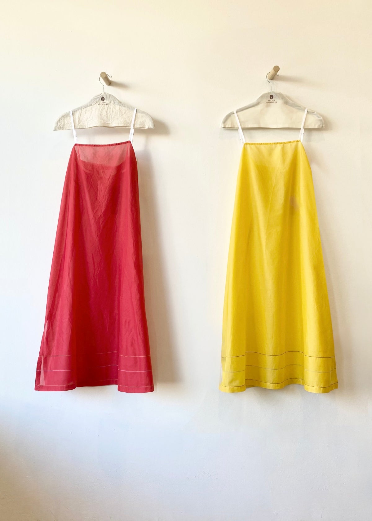 Two dresses, one red and one yellow, hanging on hangers against a white wall.