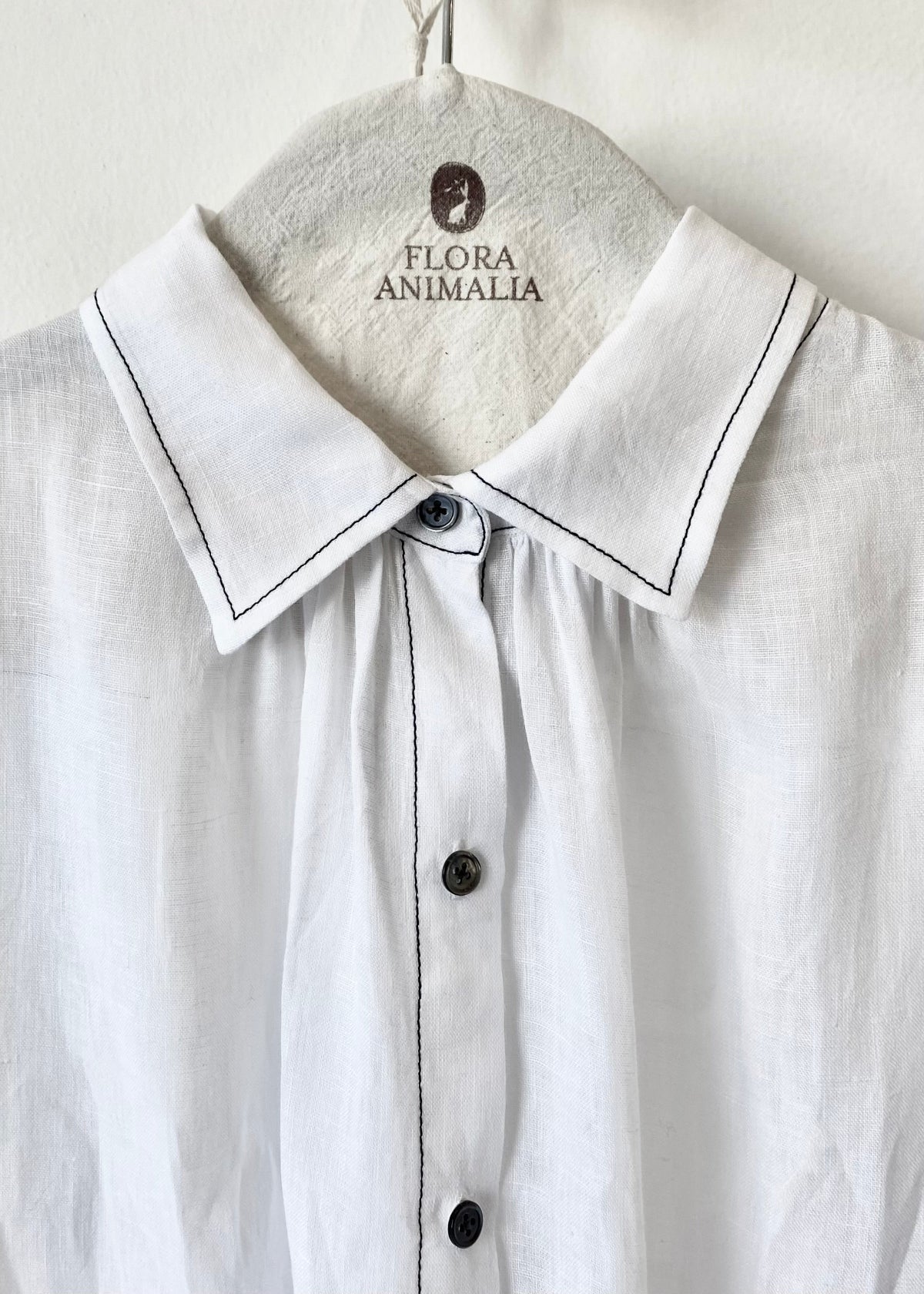 closeup detail White shirt collar with black stitching on a hanger with 'Flora Animalia' branding.