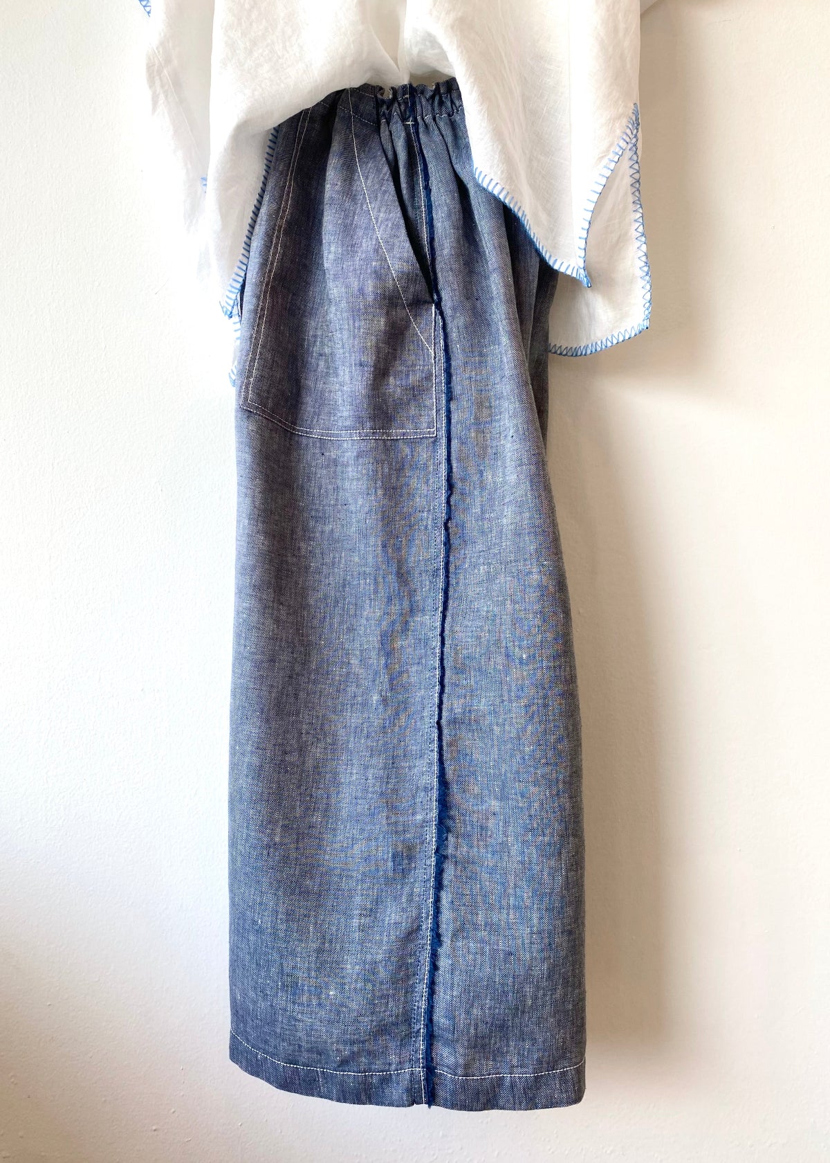 side view of blue linen pants with raw edge seam on a hanger against white wall
