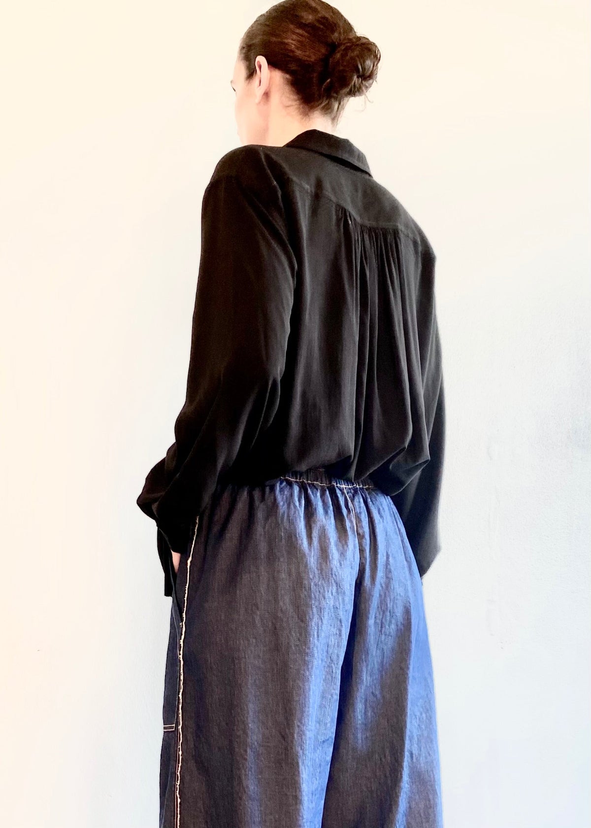 rear view of Person wearing a black Agnes shirt with back pleated yoke and blue jeans against a plain background
