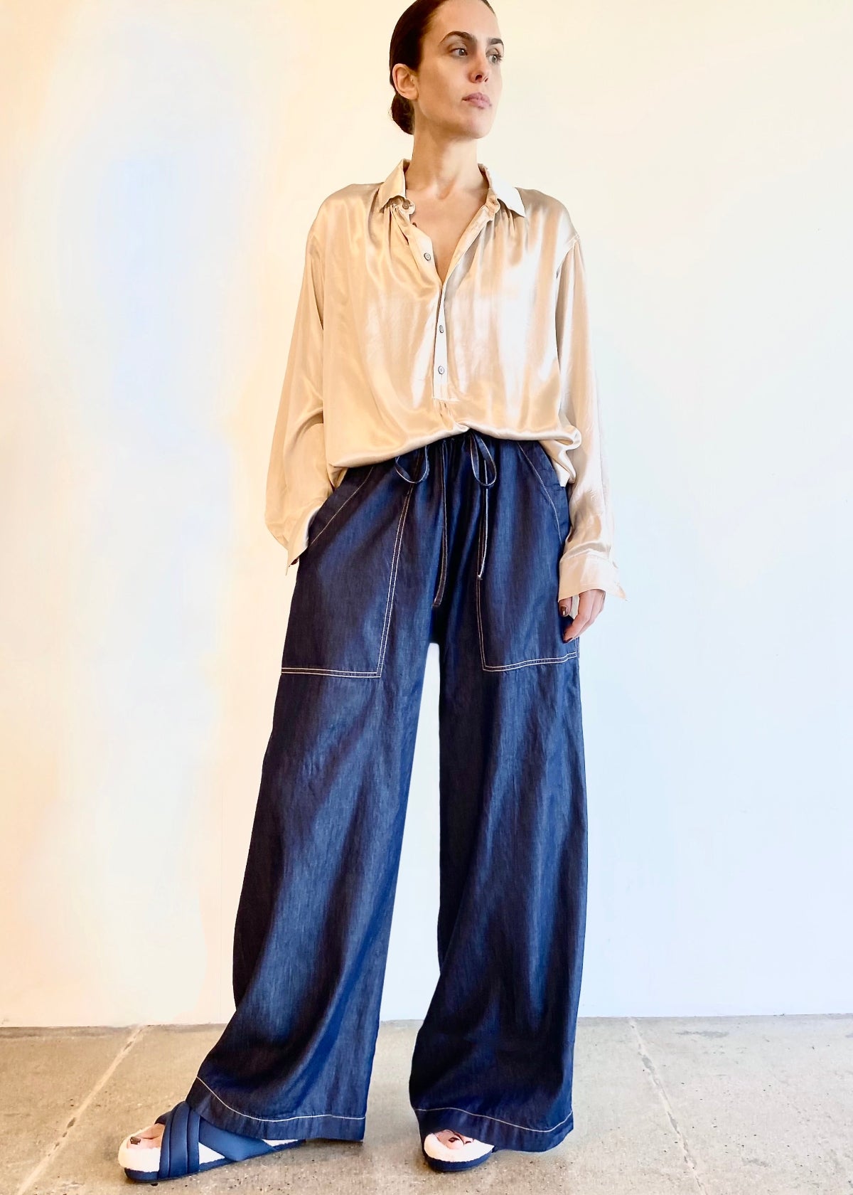 Woman wearing a beige silky blouse and blue denim wide-leg draw string pants against a plain background