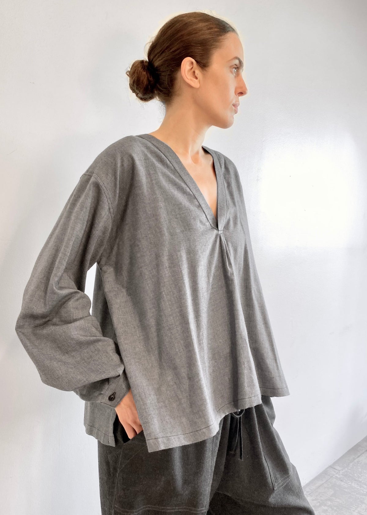 Woman wearing a light gray Farmer's Smock with dark gray drawstring flannel pants against a plain background