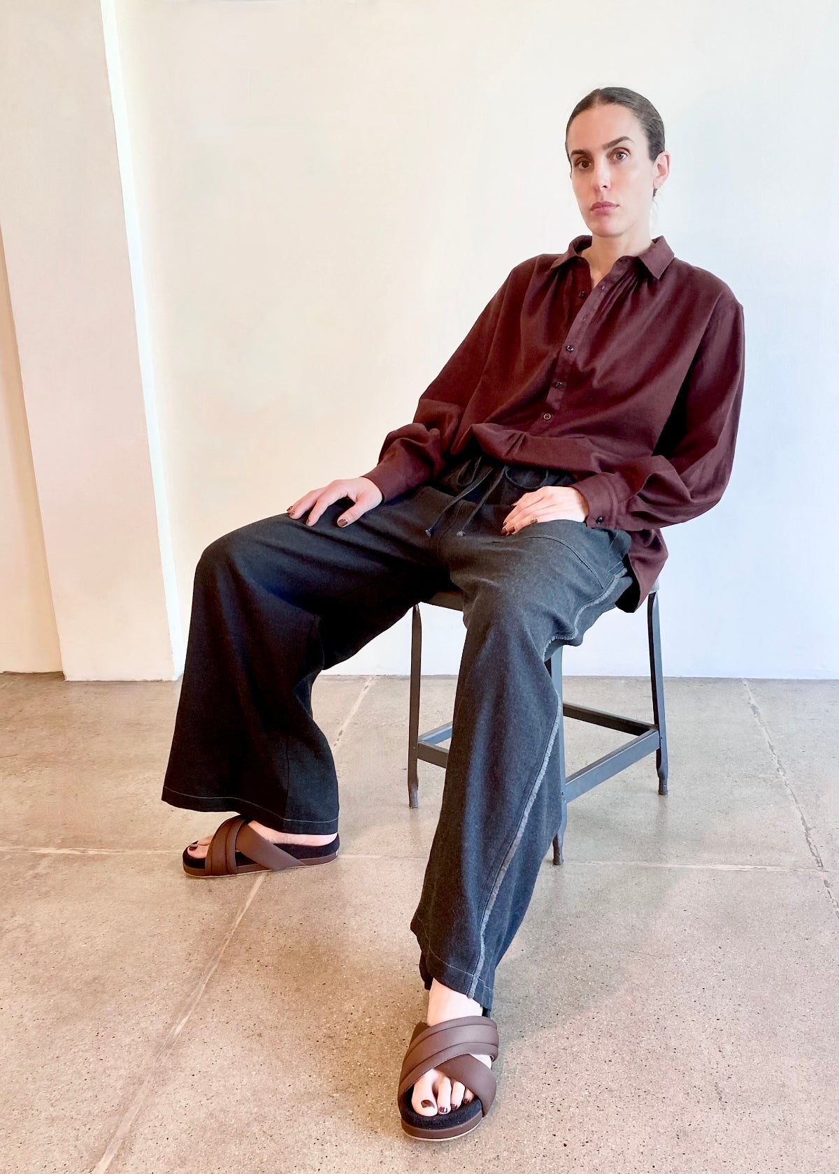 Person wearing a wine button shirt and dark pants sitting on a chair against a plain wall.
