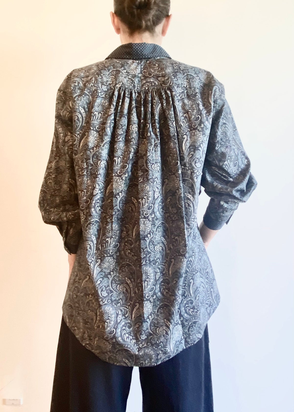 rear view of Person wearing a paisley patterned Agnes shirt and dark gray pants on a neutral background
