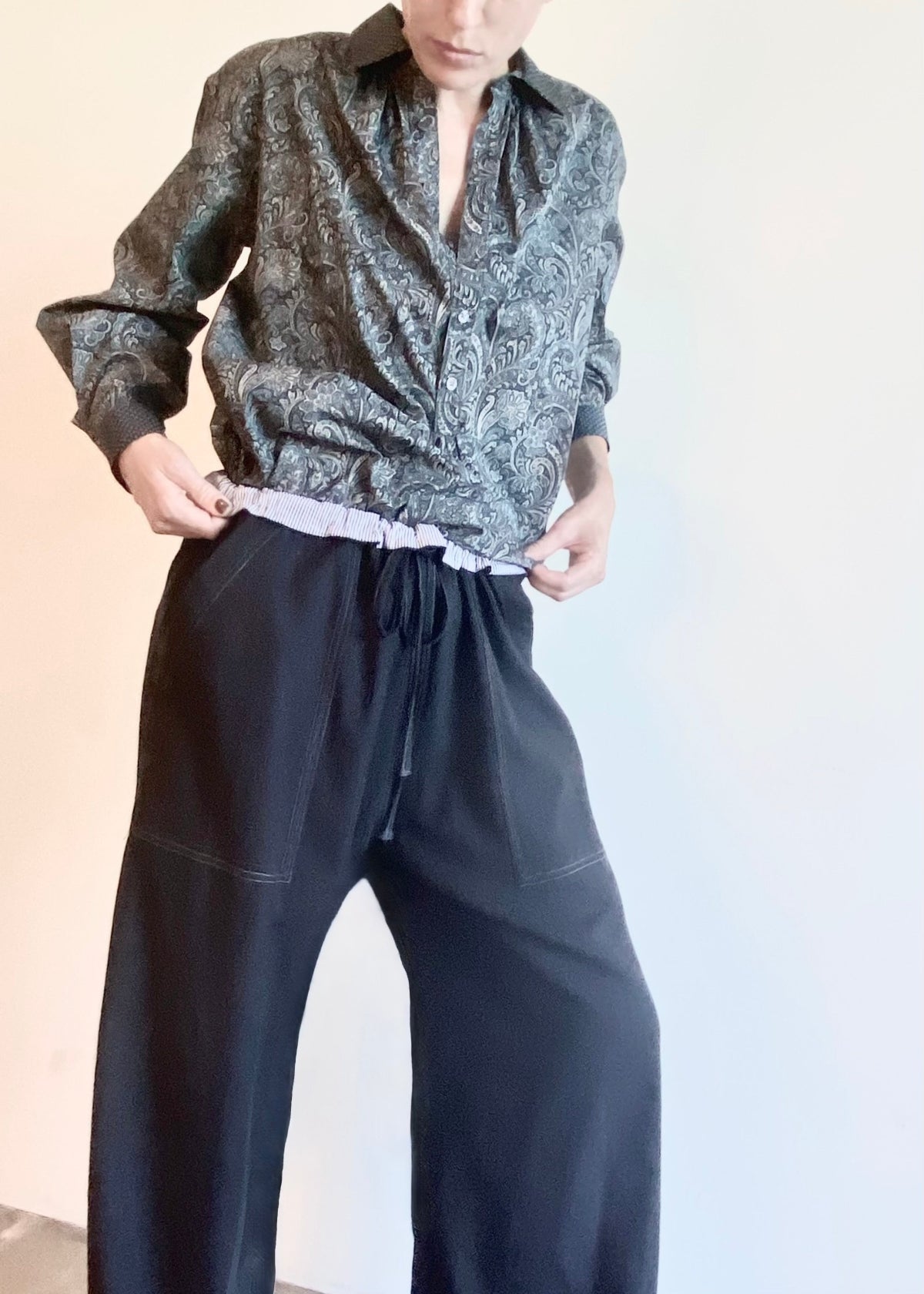 Person wearing a patterned shirt and dark pants with a rolled waistband against a plain background