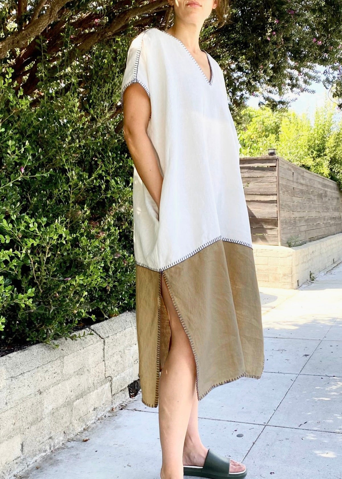 Woman wearing a white and beige dress standing outdoors with greenery in the background