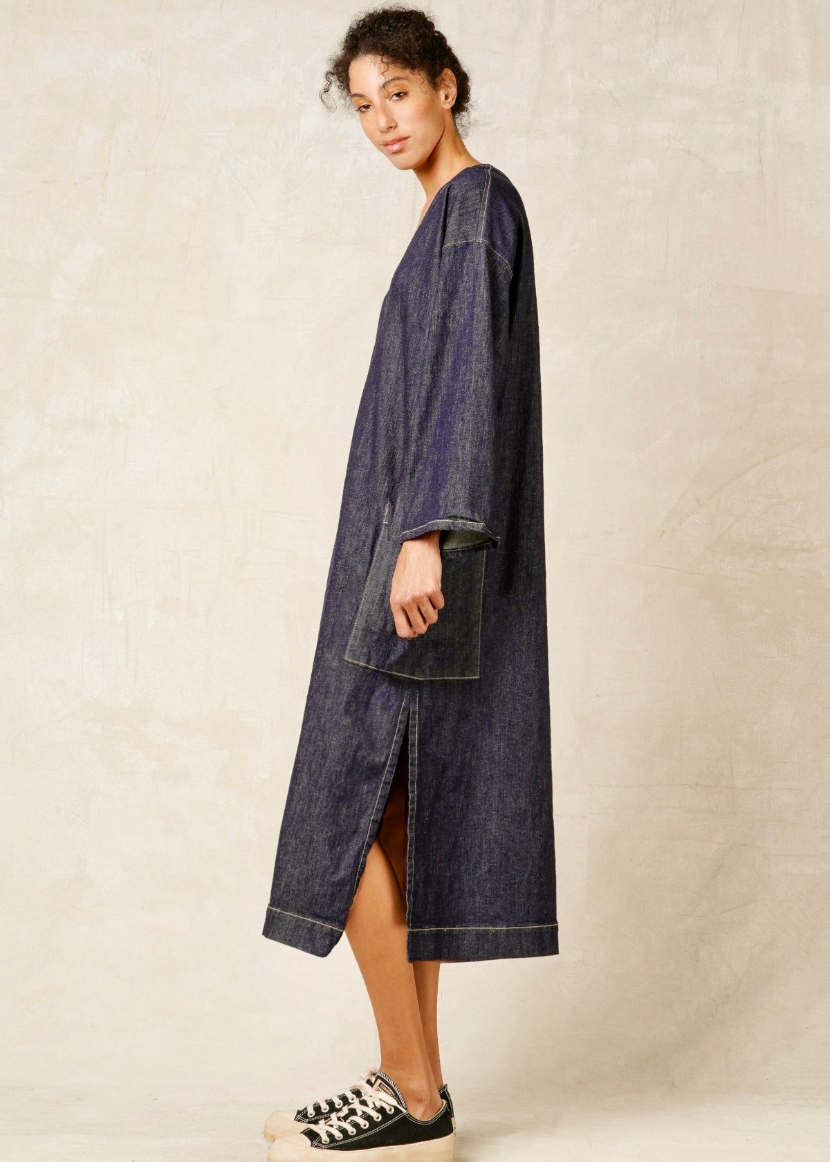 woman in v-neck loose fit denim dress with rolled sleeves and sneakers against neutral wall