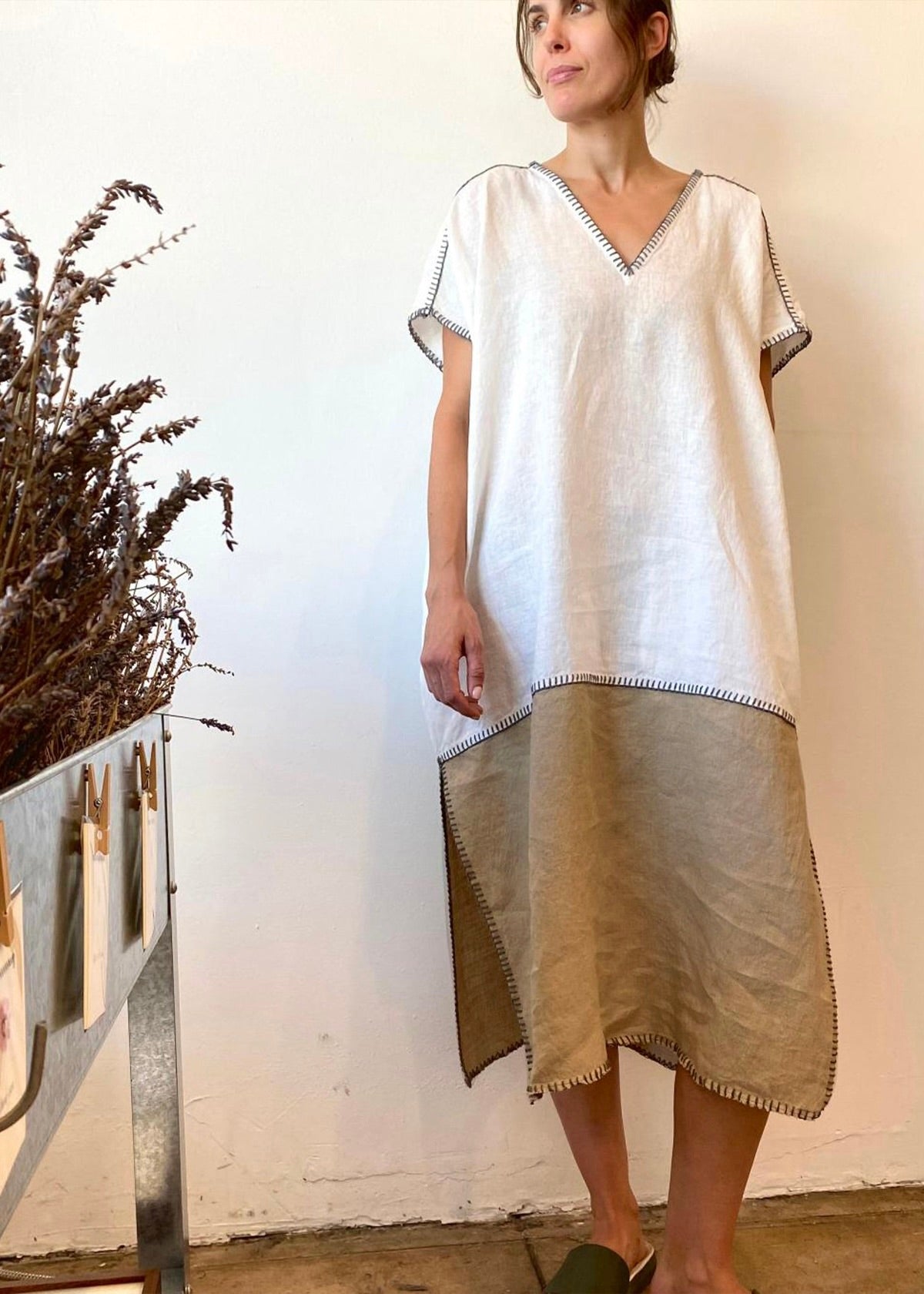 Woman wearing a white and olive dress standing against a plain wall.