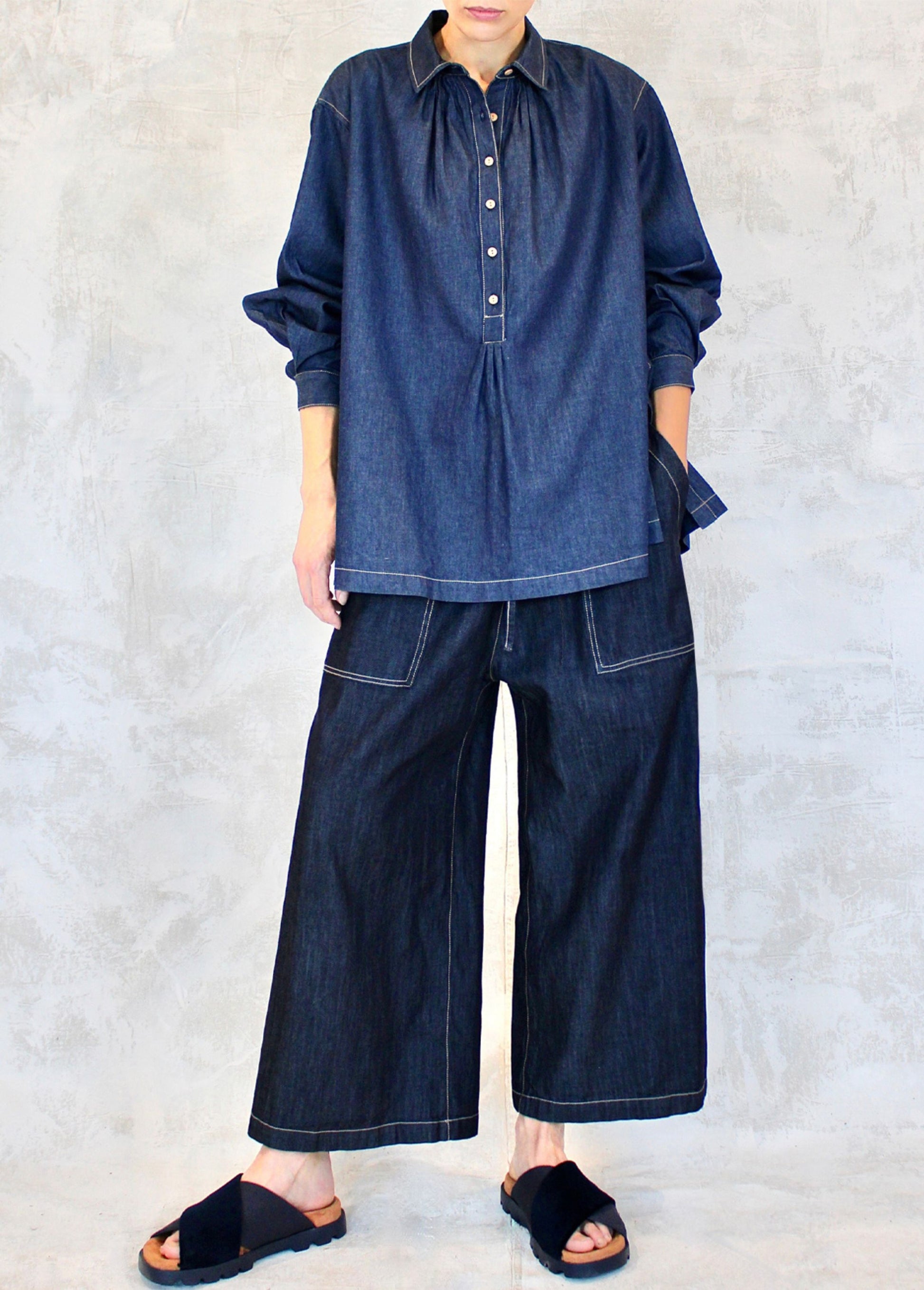 A person wearing a long-sleeved, button-up denim shirt with a classic collar, in a blue color with denim wide legged pants against a white background.