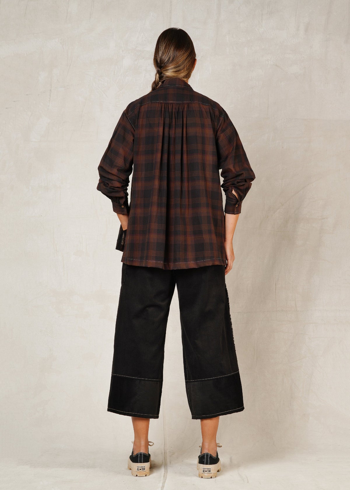 rear view of model standing on a plain background, wearing a long-sleeved checkered shirt with a collar and high-low hem, paired with black trousers and casual shoes.