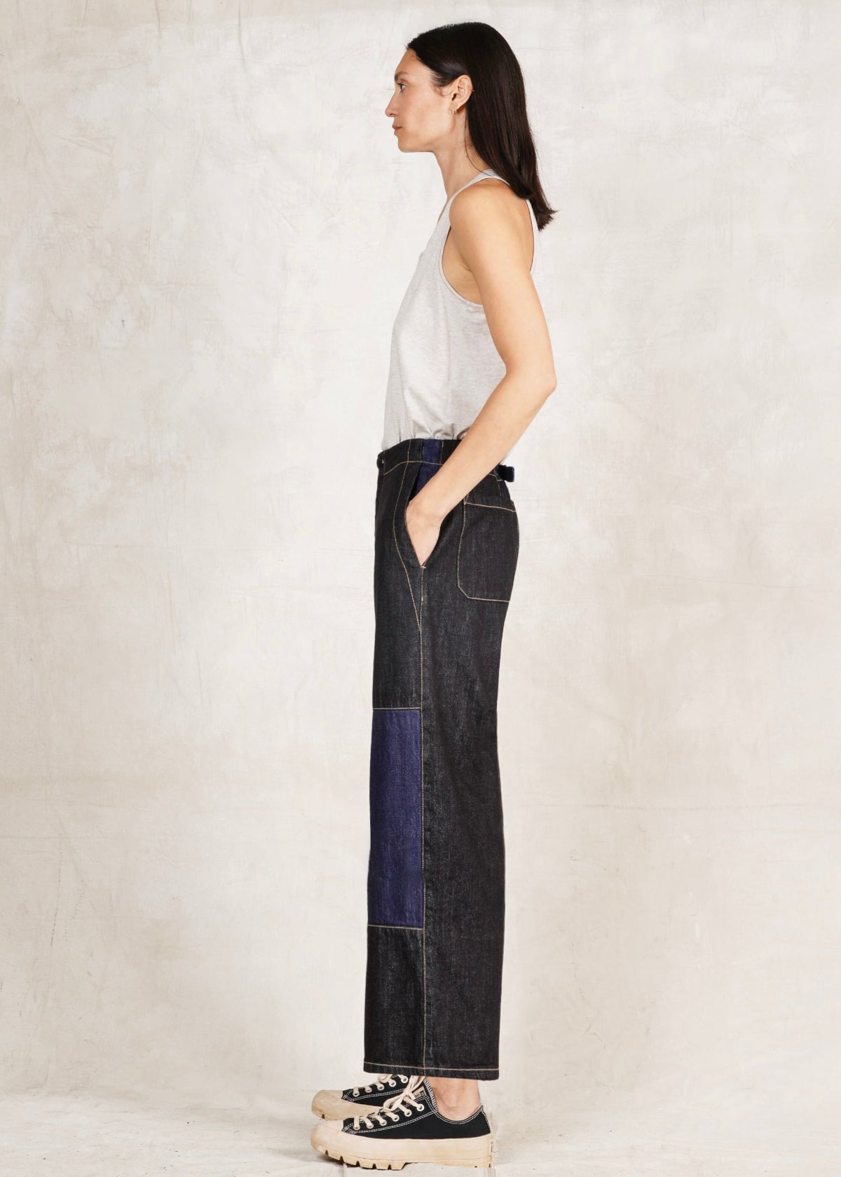A female model wearing a white sleeveless top and blue denim artist pants with a high waist cut. She is standing sideways against a plain background.