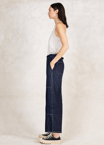 side view of Woman wearing a white top and dark blue Artist Pant jeans against a plain background