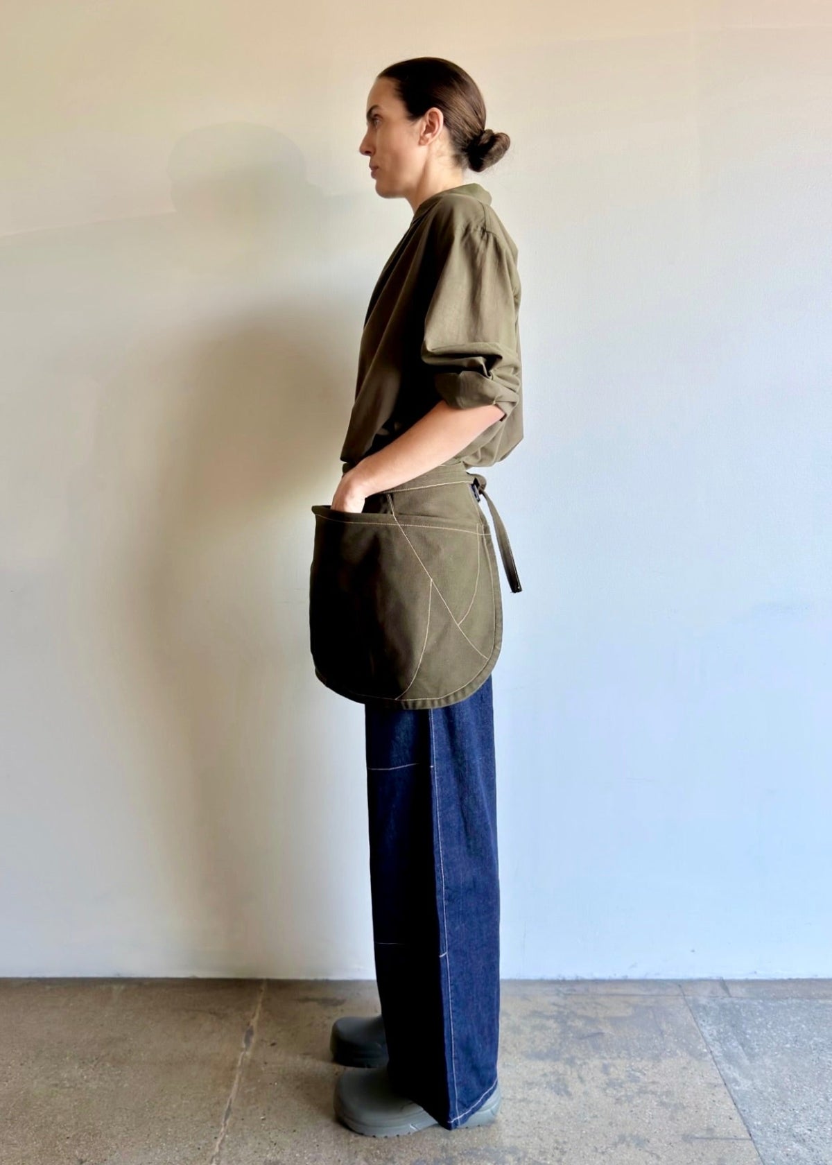 Model wearing Flora Animalia organic canvas basket apron in olive green, hand in pocket, side view.