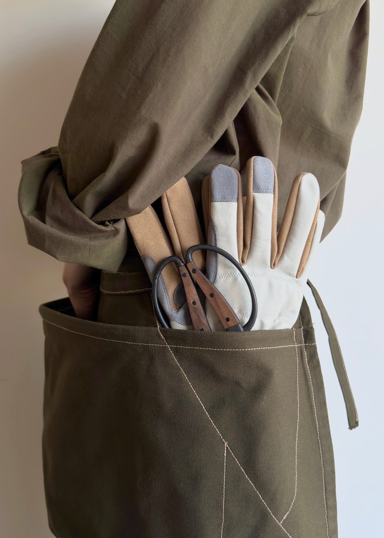 Model wearing Flora Animalia organic canvas basket apron in olive green, pocket detail with tools.