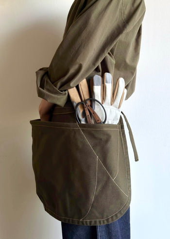 Model wearing Flora Animalia organic canvas basket apron in olive green, side view with tools in pocket.