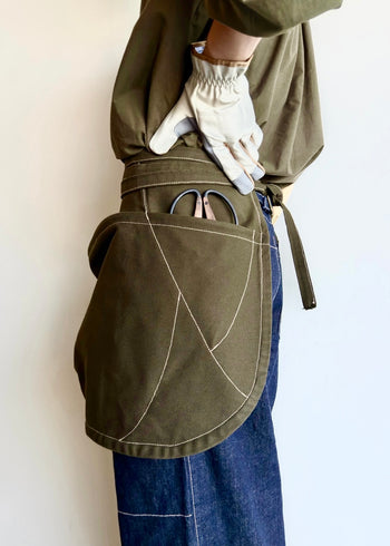 Model wearing Flora Animalia organic canvas basket apron in olive green, side view with scissors in pocket.