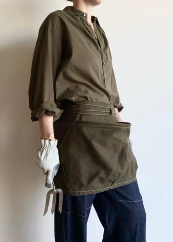 Person wearing an olive green basket apron over a shirt, holding gardening tools against a plain background