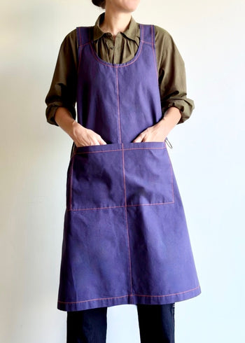 Model wearing Flora Animalia organic canvas harvest apron in purple grape, hands in pockets, front.