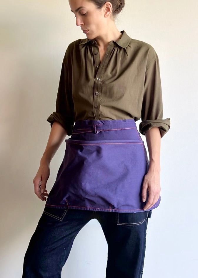 Person wearing an olive green basket apron over a shirt, holding gardening tools against a plain background