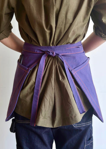 Model wearing Flora Animalia organic canvas basket apron in purple grape, back view, waist-tie detail.