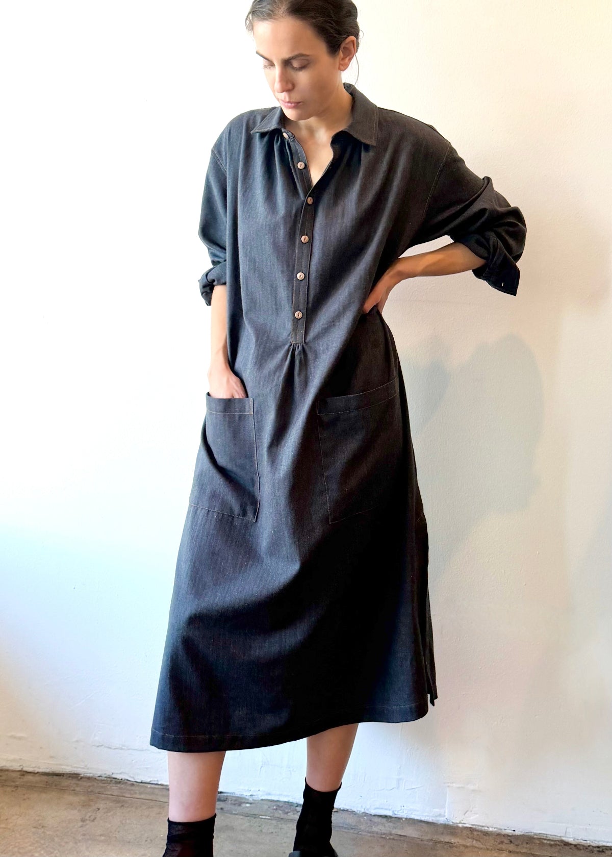 Woman wearing a long dark blue button dress with hand in pocket against a white wall.