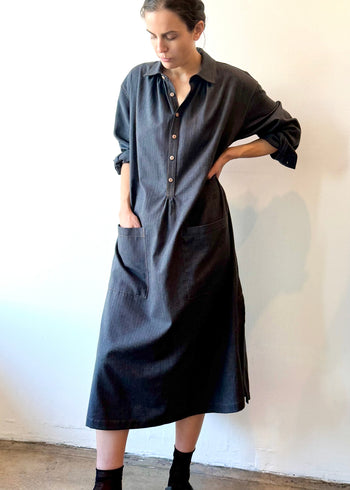 Woman wearing a long dark blue button dress with hand in pocket against a white wall.