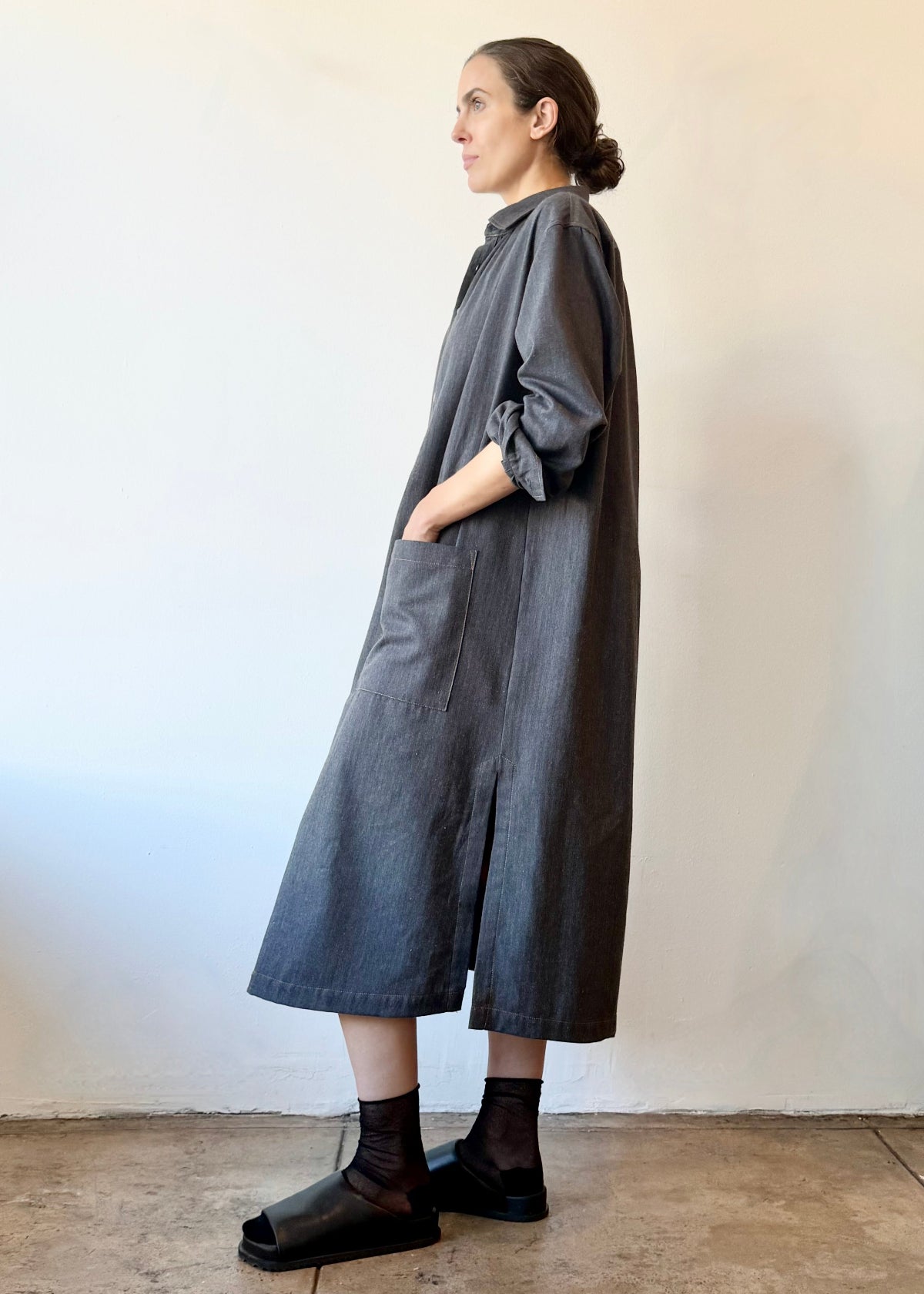 Woman wearing a long dark gray dress with hands in pockets against a plain white wall.