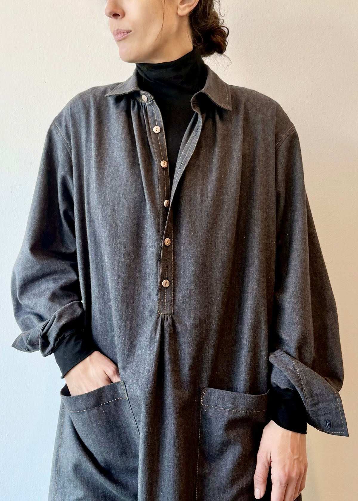 Person wearing a long dark blue button dress with black turleneck underneath  and rolled sleeves against a plain background
