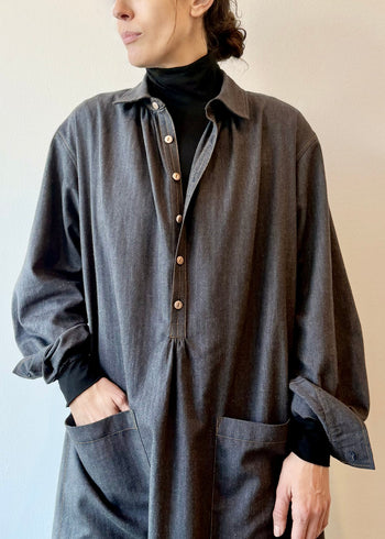 Person wearing a long dark blue button dress with black turleneck underneath  and rolled sleeves against a plain background