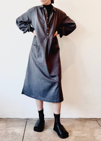 Person wearing a blue flannel button dress  with black turtleneck underneath and hands on hips against a white wall.