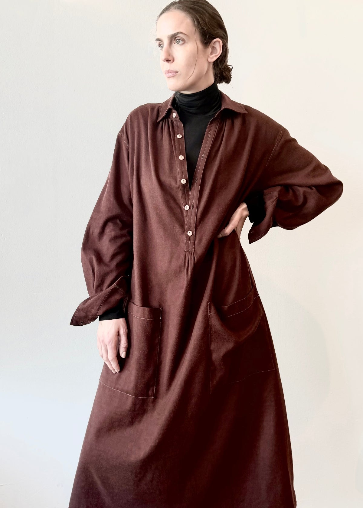 Woman wearing a long dark blue button dress with hand in pocket against a white wall.