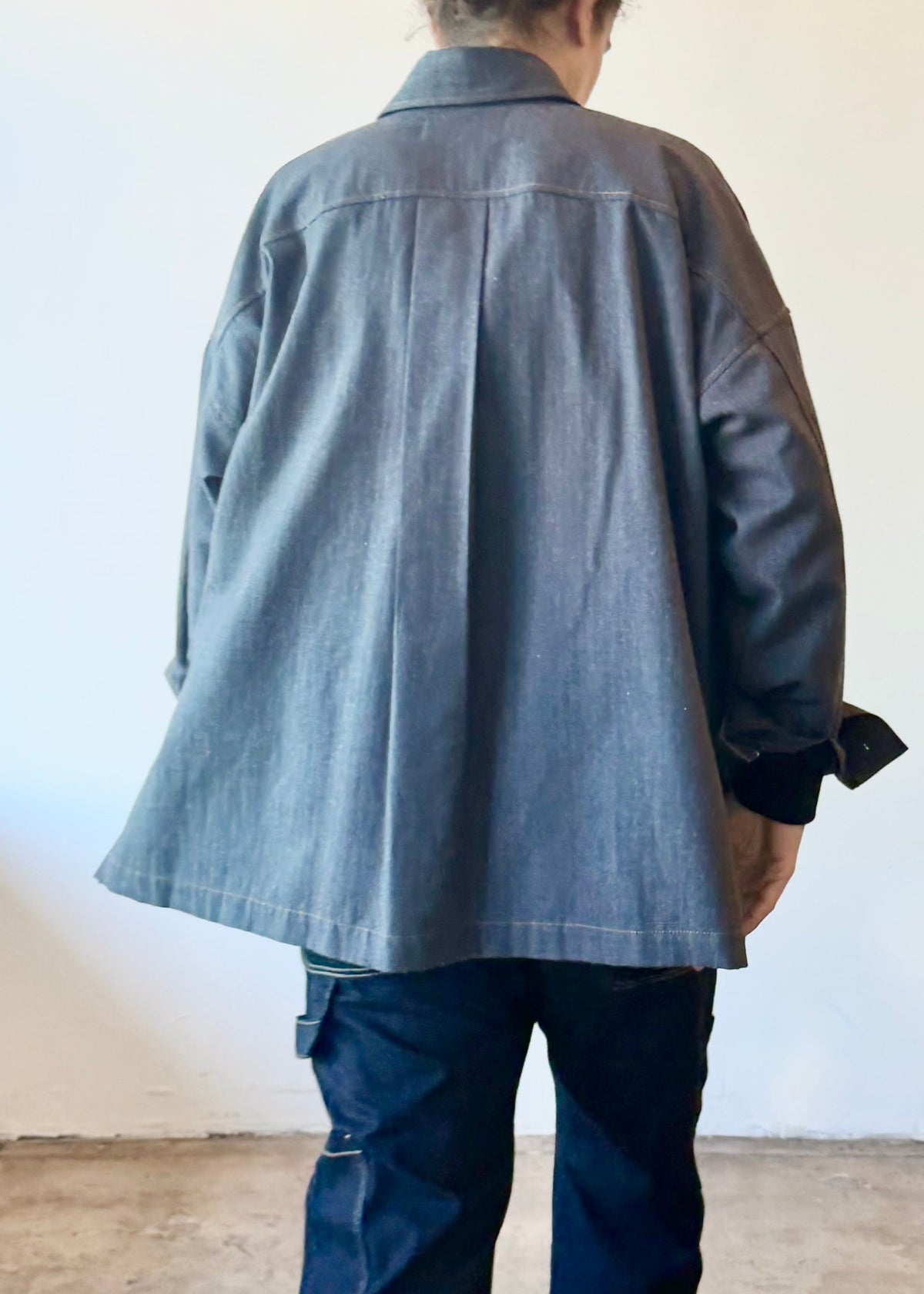 Rear of a person wearing a sleel blue colored flannel top and dark denim pants standing in a minimalistic room.