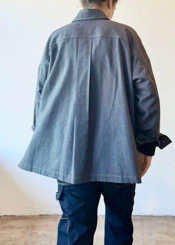 Rear of a person wearing a sleel blue colored flannel top and dark denim pants standing in a minimalistic room.