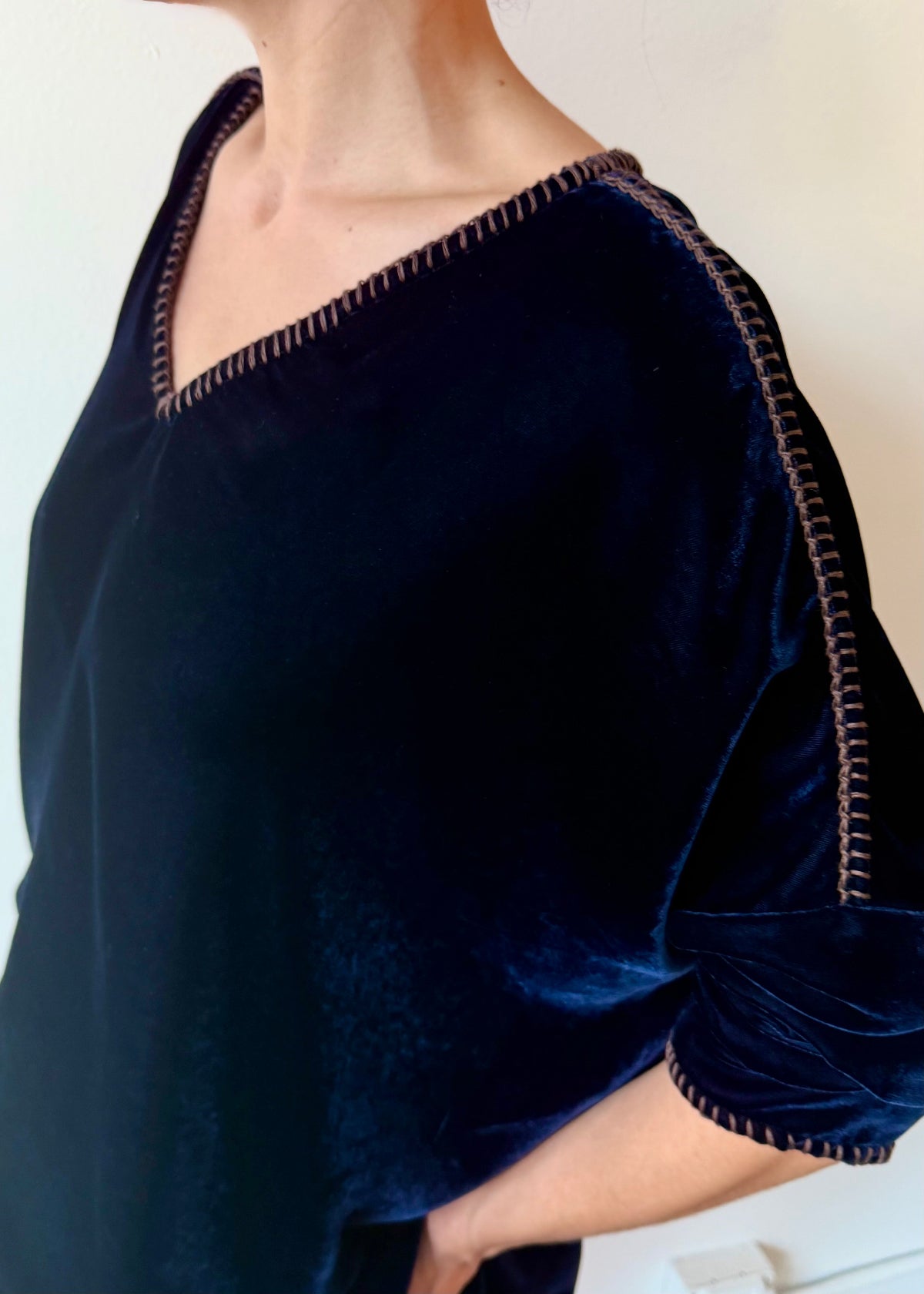 Close up of models shoulder and v-neck in a navy blue velvet top with contrast stitching detailing on a plain background.