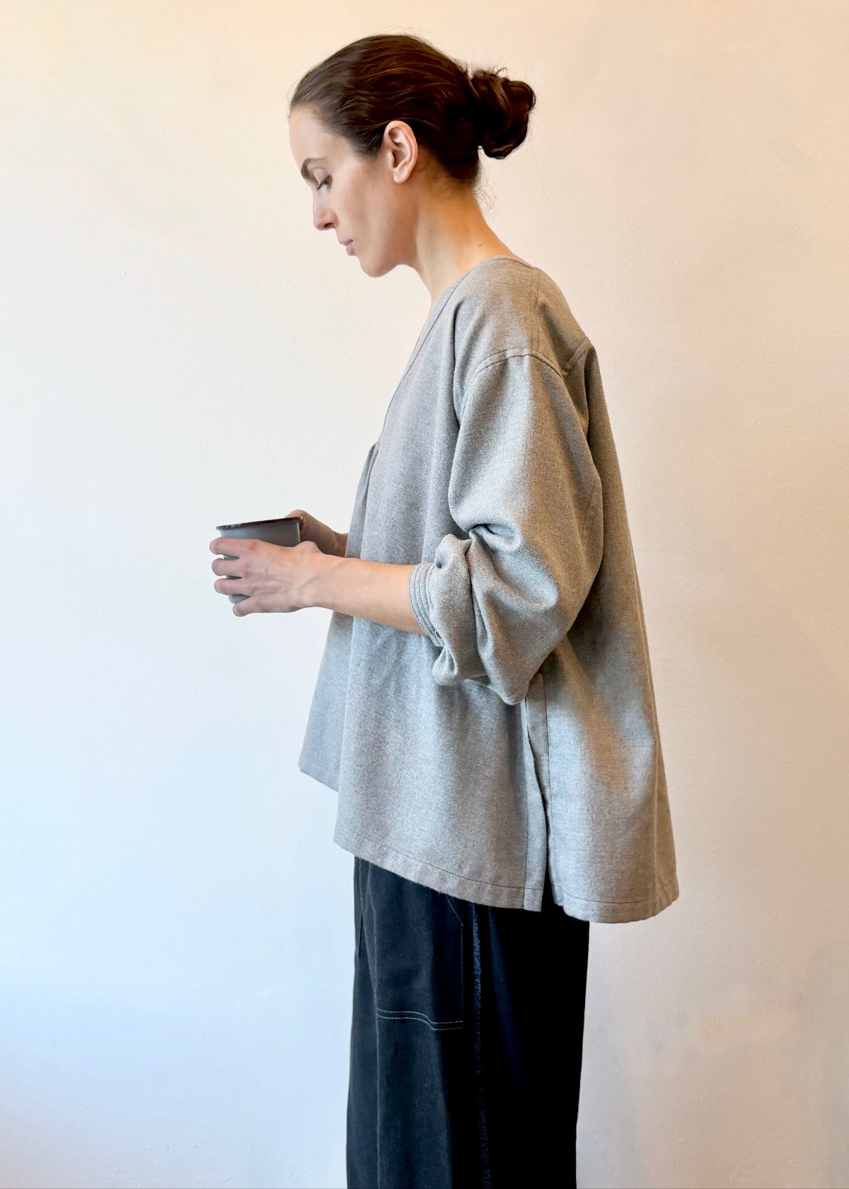 Profile view of a woman wearing a gray oversized shirt with dark flannel pants holding a cup against a plain background