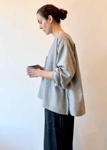 Profile view of a woman wearing a gray oversized shirt with dark flannel pants holding a cup against a plain background