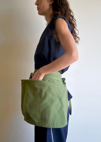 Model wearing Flora Animalia organic canvas basket apron in grass green, with hand in pocket, side view.