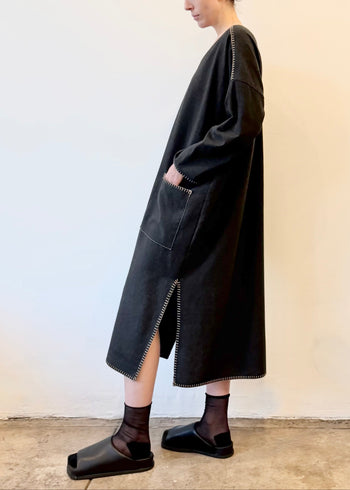 Woman wearing a dark gray caftan dress with hand in pocket standing against a plain wall.