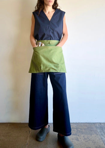 Model wearing Flora Animalia organic canvas basket apron in grass green, with hands in pocket, front view.