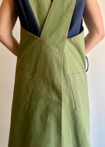 Model wearing Flora Animalia organic canvas harvest apron in grass green, back view showing cross-back straps, pockets and utility ring.