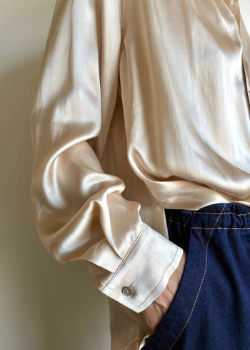 Close-up of a neutral biosilk Agnes shirt with button sleeve and a drape tuck, worn with dark denim pants against a neutral background.