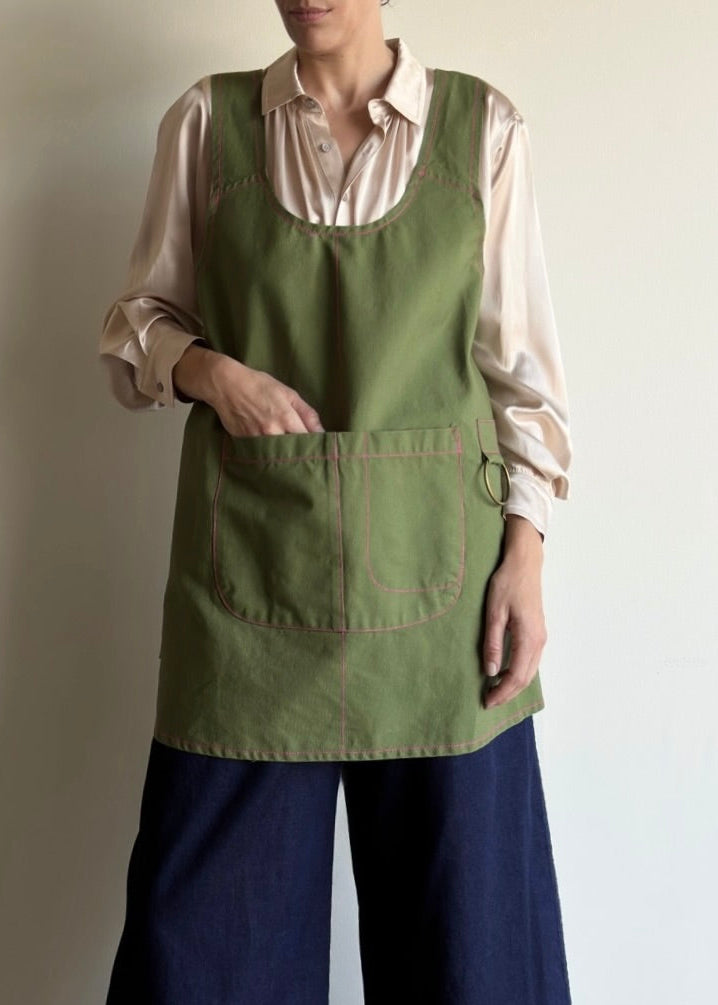 Woman wearing a grass green apronette over a light shirt and dark pants against a plain background