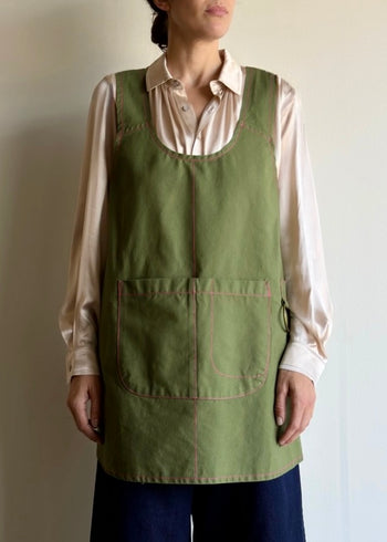 Person wearing a grass green apronette apron over a light-colored shirt against a plain background