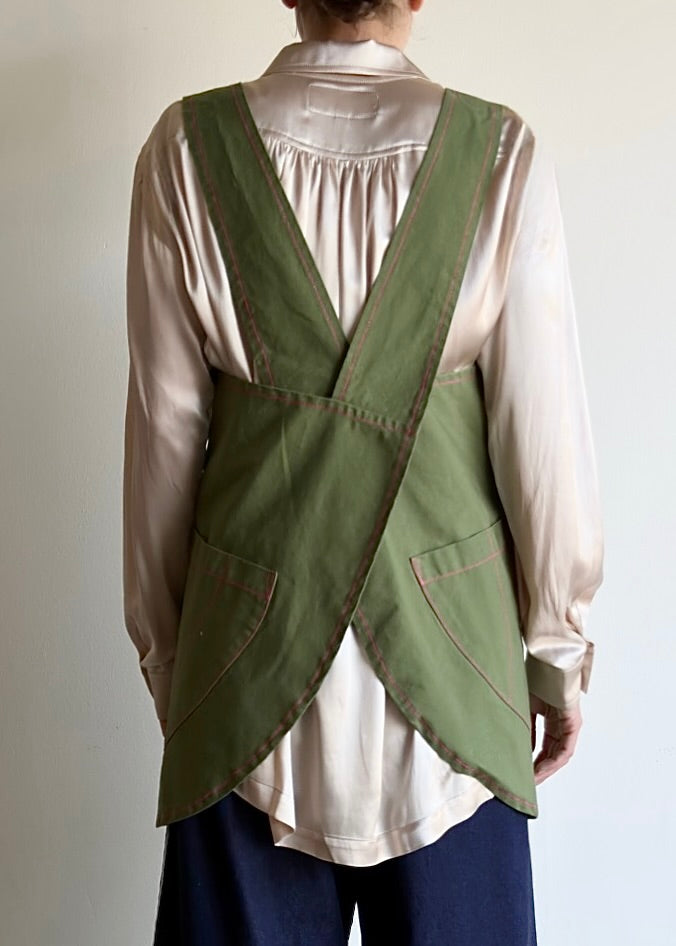 Cross straps, rear view of Person wearing a green apronette over a pink shirt with a neutral background