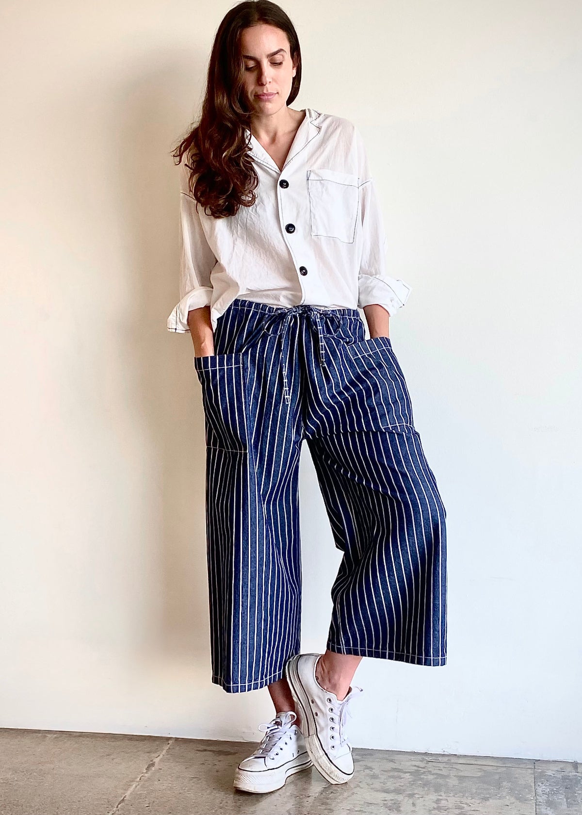 blue and white striped denim cropped drawstring pants with white button down top. Hands in pockets