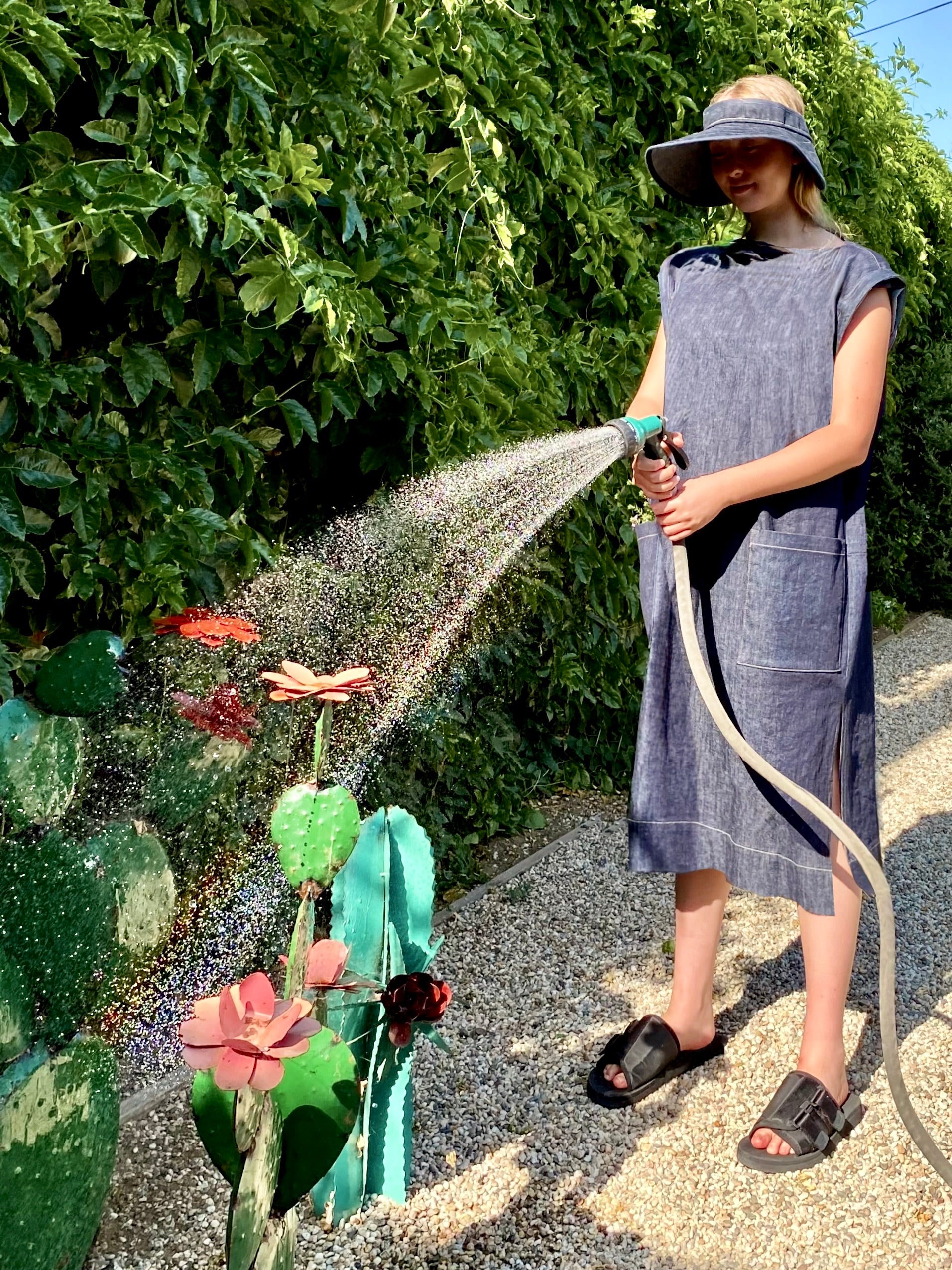 Person in a denim dress and visor watering plants with a hose in a garden setting