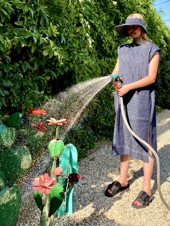 Person in a denim dress and visor watering plants with a hose in a garden setting