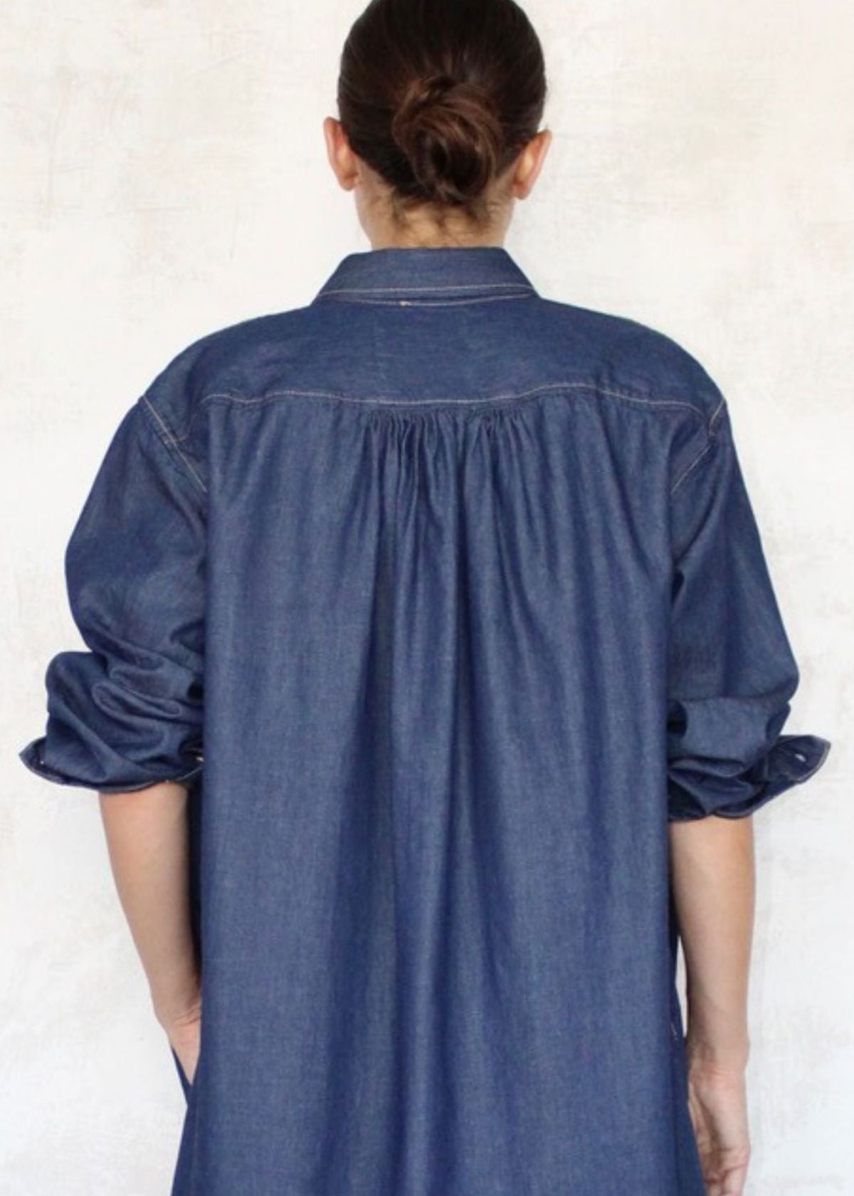 Rear close up of person wearing a blue denim Agnes dress with rolled-up sleeves on a white background