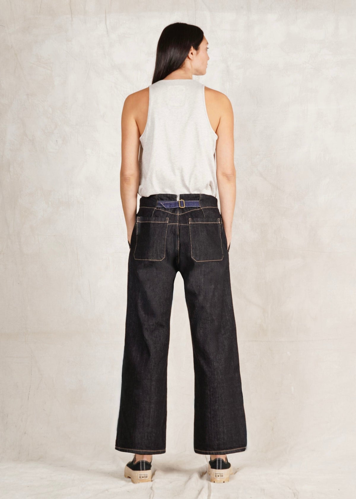 A female model wearing a white sleeveless top and blue denim artist pants with a high waist cut. She is standing against a plain background with back to us